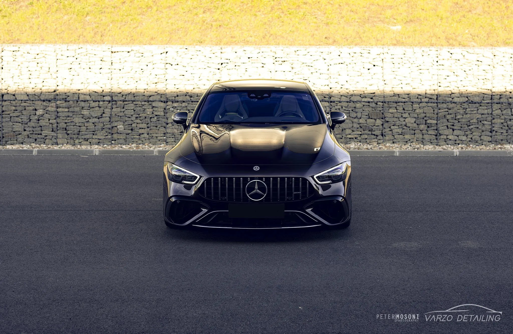 Barna Mercedes-AMG GT négyajtós kupé szemből, kőfal előtt